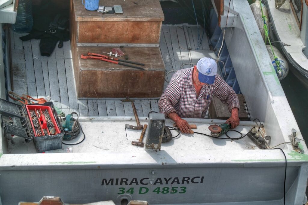 A man repairs equipment on a fishing boat at a dock, surrounded by tools and gear.